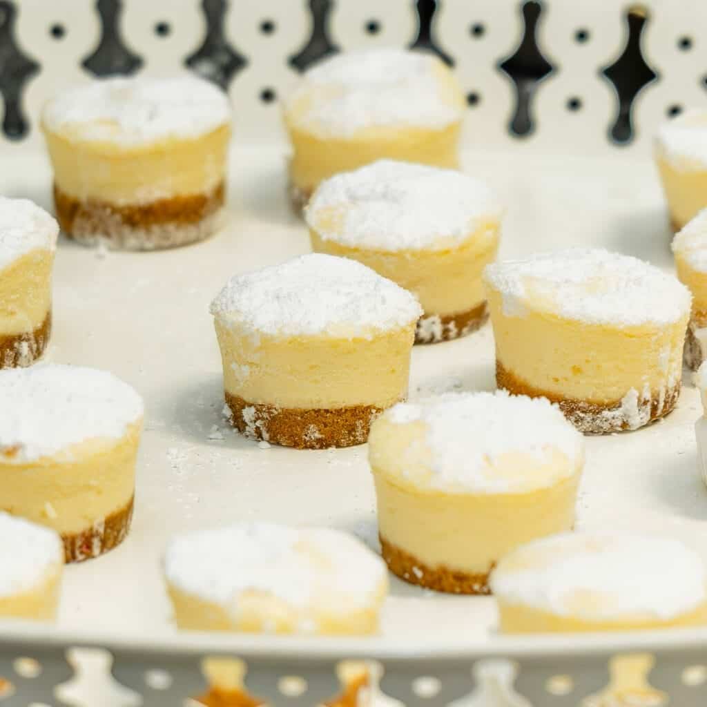 Creamy lemon cheesecakes with a biscuit base, dusted with powdered sugar, on a decorative tray.