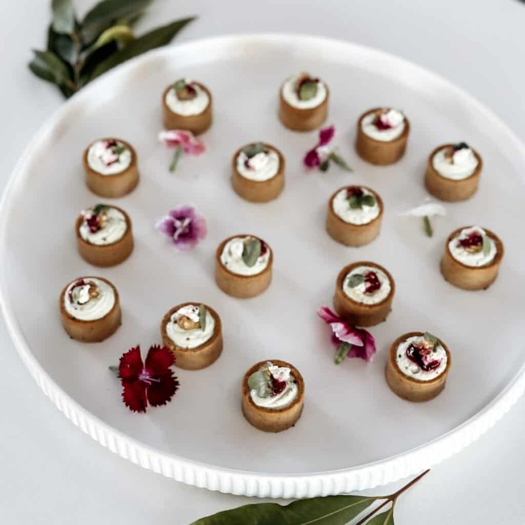 Miniature savory tartlets with cream and herbs arranged on a white platter, garnished with edible fl.